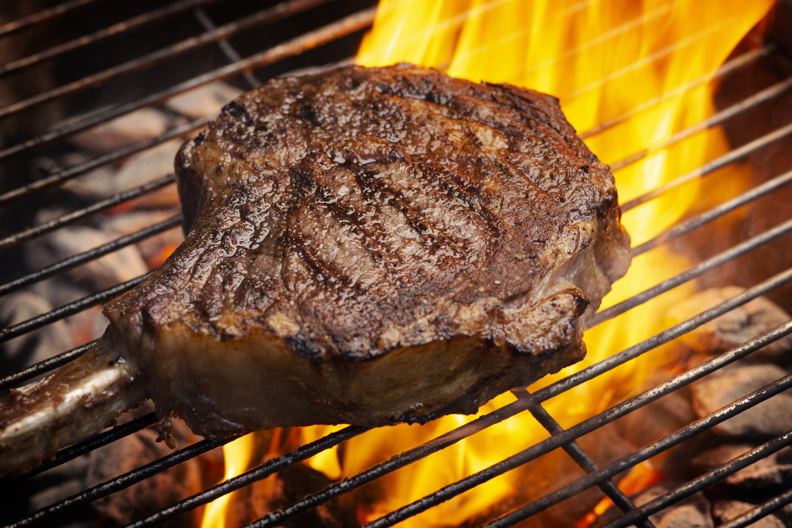 a bone-in tomahawk ribeye steak cooking on grill with flames, angled view